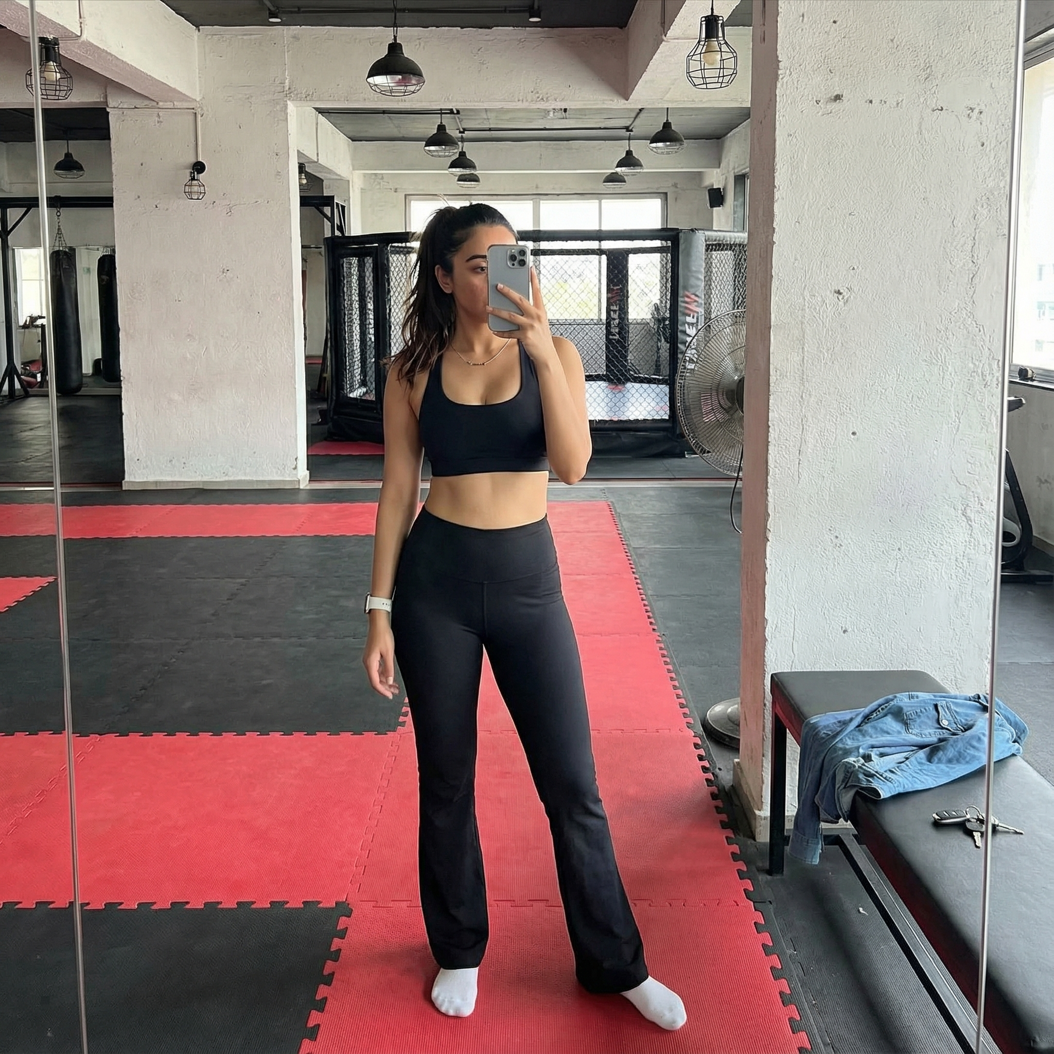 {
  "subject": {
    "description": "The person from the input image taking a full-body mirror selfie while standing on gym mats",
    "age": "match the person's real age",
    "expression": "face partially or fully obscured by the phone",
    "hair": {
      "color": "match the person's natural hair color",
      "style": "high ponytail with optional loose face-framing strands (adapt naturally to the person's hair type)"
    },
    "clothing": {
      "top": {
        "type": "athletic sports bra or fitted athletic top",
        "color": "black",
        "details": "minimal, scoop neck, tight athletic fit"
      },
      "bottom": {
        "type": "flared athletic yoga pants",
        "color": "black",
        "details": "high-waisted, fitted through thighs and slightly flared at the hem, paired with white ankle socks"
      }
    },
    "face": {
      "preserve_original": true,
      "makeup": "unnecessary or minimal since the face is covered by the phone"
    }
  },
  "accessories": {
    "jewelry": {
      "wrist": "white smartwatch band on the left wrist",
      "neck": "thin delicate chain necklace"
    },
    "device": {
      "type": "smartphone",
      "details": "silver or light-colored phone held vertically, covering part or all of the face"
    }
  },
  "photography": {
    "camera_style": "modern smartphone mirror selfie aesthetic",
    "angle": "standing eye-level reflection",
    "shot_type": "full-body vertical shot, 9:16 aspect ratio",
    "texture": "sharp focus, realistic textures, natural daylight contrast, soft shadows"
  },
  "background": {
    "setting": "industrial MMA or combat-style gym interior",
    "wall_color": "white exposed concrete",
    "elements": [
      "interlocking red and black foam gym mats on the floor",
      "black industrial pendant cage lights hanging overhead",
      "exposed white concrete ceiling with visible pipes",
      "large black industrial fan to the left",
      "wooden slat bench in the foreground",
      "black textured foam roller on the floor",
      "crumpled blue denim shirt and keys on the bench",
      "blurred MMA cage and chain-link fencing in the background"
    ],
    "atmosphere": "authentic training environment, gritty but bright",
    "lighting": "mix of natural daylight from large windows and overhead gym lights"
  }
}