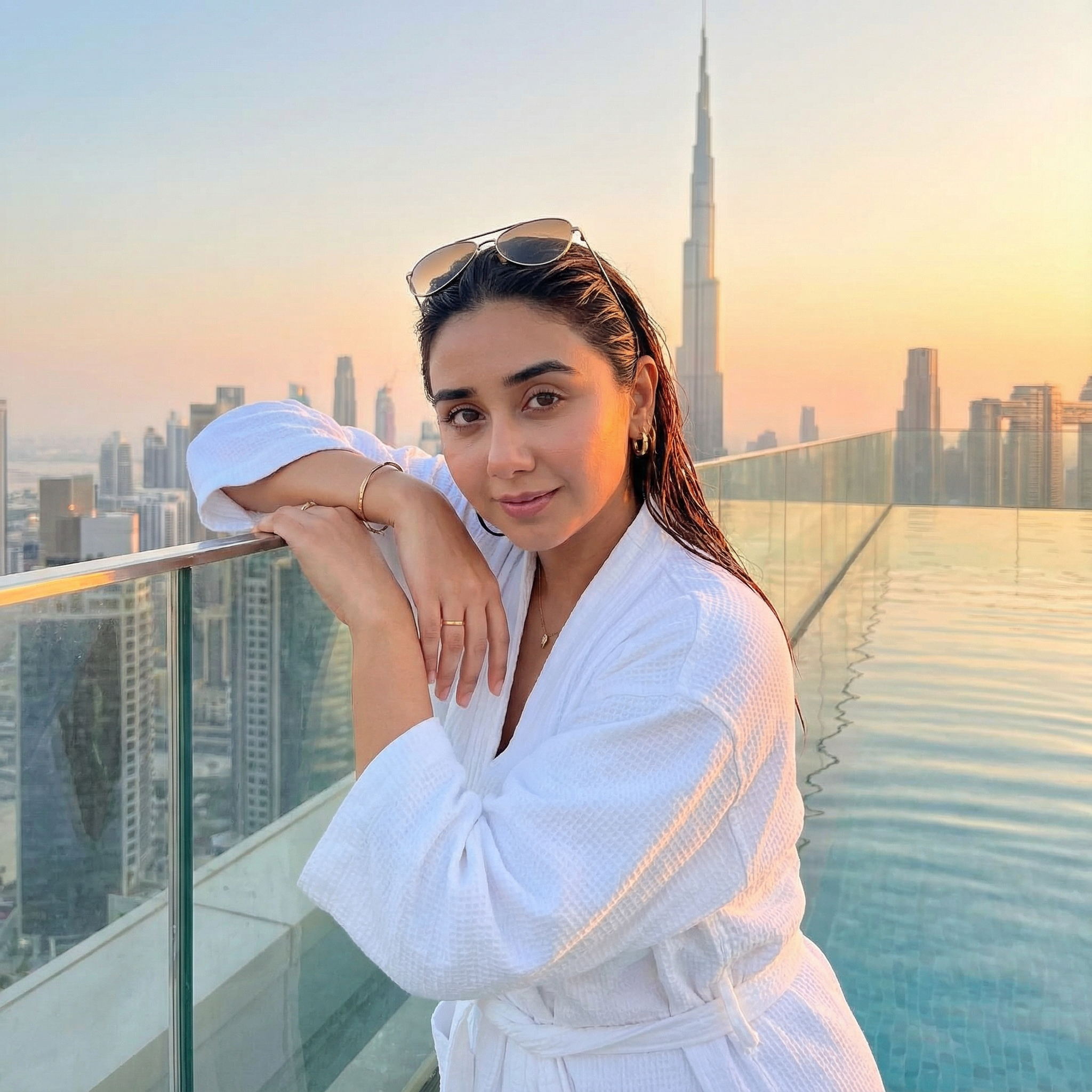 {
  "subject": {
    "description": "The person from the input image resting their arms on the glass edge of a rooftop infinity pool, overlooking the Dubai skyline during golden hour",
    "expression": "serene, relaxed, soft smile with dreamy eyes",
    "hair": {
      "style": "wet or slicked-back look to match pool environment",
      "details": "sunlight catching tiny droplets"
    },
    "clothing": {
      "outfit": "premium minimalist swimwear or luxurious white resort robe draped around shoulders",
      "details": "skin glowing with warm sunlight, soft reflections from water"
    }
  },
  "accessories": {
    "eyewear": "luxury aviator sunglasses placed on head or worn",
    "jewelry": "water-safe minimal gold jewelry"
  },
  "photography": {
    "camera_style": "luxury travel influencer aesthetic",
    "angle": "eye-level but slightly tilted for cinematic depth",
    "shot_type": "waist-up pool portrait",
    "texture": "smooth reflections, golden highlights, clean glossy water ripples"
  },
  "background": {
    "setting": "Dubai skyscraper skyline",
    "elements": [
      "Burj Khalifa in far distance",
      "sunset gradient from orange to soft rose",
      "clear blue infinity pool water merging with skyline",
      "glass railing reflecting warm tones"
    ],
    "atmosphere": "ultra-rich vacation serenity",
    "lighting": "golden hour glow with soft shadows"
  }
}