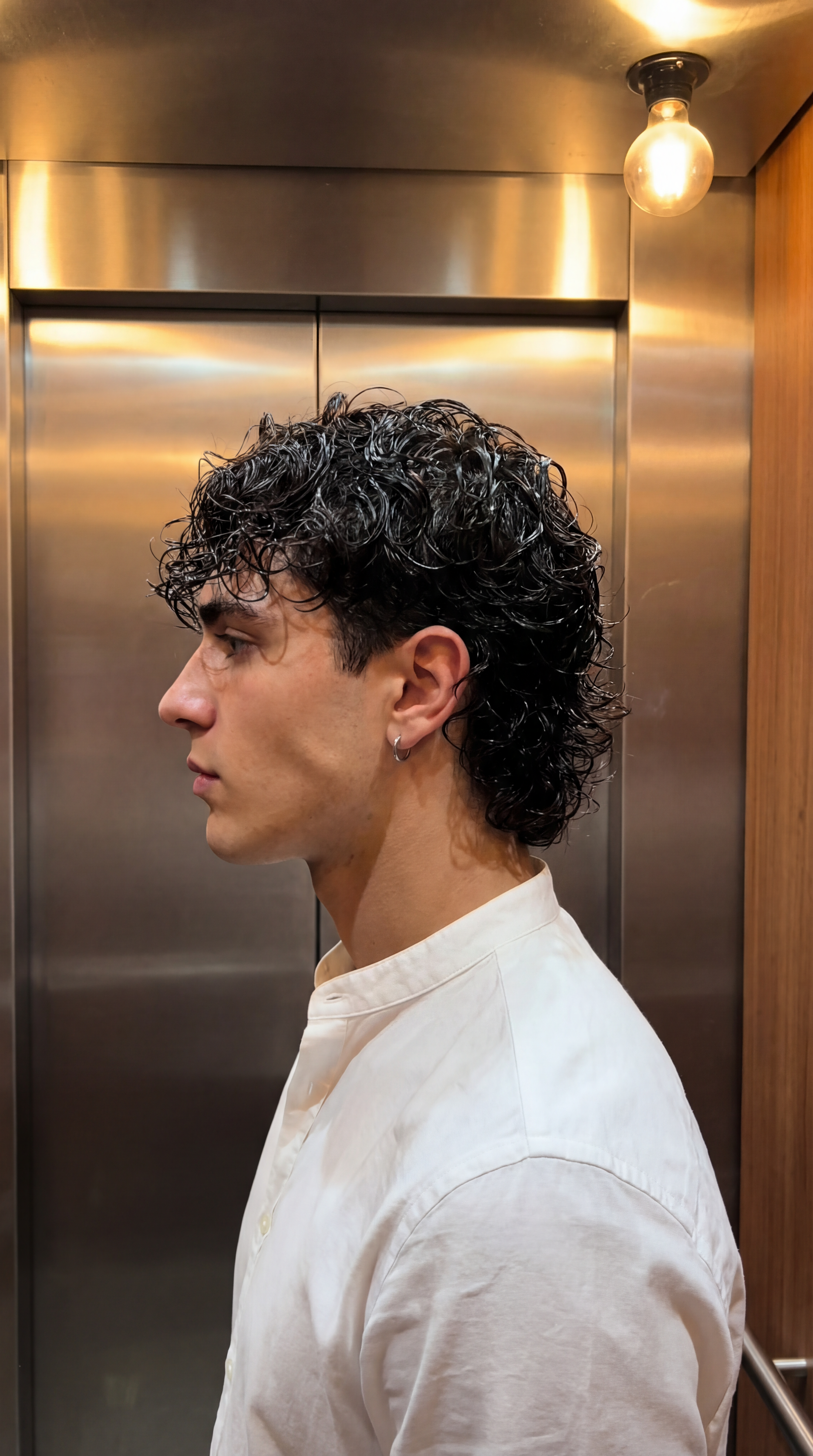 { "metadata": { "confidence_score": "high", "image_type": "photograph", "primary_purpose": "social media/portrait" }, "composition": { "rule_applied": "center composition", "aspect_ratio": "9:16 vertical", "layout": "single subject", "focal_points": [ "Subject's profile and hair texture", "Exposed light bulb in upper right" ], "visual_hierarchy": "Eye is drawn to the high-contrast lighting on the hair, then down the profile face, then to the background environment", "balance": "asymmetric - subject faces left into negative space, balanced by light fixture on right" }, "color_profile": { "dominant_colors": [ { "color": "Brushed Metal Grey", "hex": "#8C8B88", "percentage": "40%", "role": "background" }, { "color": "Warm Wood Brown", "hex": "#966F46", "percentage": "20%", "role": "background accent" }, { "color": "Crisp White", "hex": "#F0F2F5", "percentage": "20%", "role": "clothing" }, { "color": "Dark Espresso Hair", "hex": "#2A1F1D", "percentage": "10%", "role": "primary subject" } ], "color_palette": "neutral with warm accents", "temperature": "warm", "saturation": "moderate", "contrast": "medium-high contrast" }, "lighting": { "type": "artificial/mixed", "source_count": "single source visible", "direction": "top-down/back-right", "directionality": "highly directional", "quality": "hard light", "intensity": "bright/high-key highlights", "contrast_ratio": "medium contrast", "mood": "candid/urban/raw", "shadows": { "type": "defined edges", "density": "gray", "placement": "under jawline/on neck/behind subject on metal doors", "length": "short" }, "highlights": { "treatment": "specular/glossy", "placement": "strong specular highlights on wet-look hair/rim light on profile" }, "ambient_fill": "present", "light_temperature": "warm (tungsten)" }, "technical_specs": { "medium": "digital photography", "style": "realistic/candid/snapshot", "texture": "smooth metallic vs organic hair", "sharpness": "tack sharp on subject", "grain": "minimal digital noise", "depth_of_field": "medium - background distinct but slightly separated", "perspective": "eye-level profile" }, "artistic_elements": { "genre": "portrait/street/lifestyle", "influences": [ "Social media aesthetic", "Urban candid photography" ], "mood": "calm/contemplative/casual", "atmosphere": "Everyday urban transition moment, quiet", "visual_style": "clean/structured" }, "typography": { "present": false, "fonts": [], "placement": "none", "integration": "none" }, "subject_analysis": { "primary_subject": "Young male adult in profile", "positioning": "center-left", "scale": "medium close-up (bust shot)", "interaction": "isolated/passive", "facial_expression": { "mouth": "neutral/closed", "smile_intensity": "no smile", "eyes": "open/gaze directed forward-left/neutral", "eyebrows": "neutral/relaxed", "overall_emotion": "serious/calm/composed", "authenticity": "candid/natural" }, "hair": { "length": "short-to-medium/nape length", "cut": "modern shag/mullet-lite/layered", "texture": "wavy/curly (Type 2C)", "texture_quality": "thick/coarse", "natural_imperfections": "flyaways at crown/natural curl clumping", "styling": "wet look/product-heavy/defined curls", "styling_detail": "Heavily styled with gel or mousse creating distinct, glossy curl definition and wet appearance", "part": "indistinct/messy", "volume": "moderate volume", "details": "Hair is longer at the nape, curling outwards; shorter layers on top and sides; glossy finish reflects overhead light strongly" }, "hands_and_gestures": { "left_hand": "not visible", "right_hand": "not visible", "finger_positions": "n/a", "finger_interlacing": "n/a", "hand_tension": "n/a", "interaction": "hands are out of frame", "naturalness": "n/a" }, "body_positioning": { "posture": "standing straight", "angle": "pure profile (90 degree turn to left)", "weight_distribution": "centered", "shoulders": "relaxed/perpendicular to camera" } }, "background": { "setting_type": "indoor/elevator interior", "spatial_depth": "shallow", "elements_detailed": [ { "item": "Elevator Doors", "position": "center/background", "distance": "background", "size": "dominant", "condition": "clean/industrial", "specific_features": "brushed metal texture, vertical seam" }, { "item": "Hanging Light Bulb", "position": "top right", "distance": "foreground/midground", "size": "small", "condition": "functional", "specific_features": "exposed bulb style, warm glow" }, { "item": "Wall Panel", "position": "right", "distance": "background", "size": "medium", "condition": "good", "specific_features": "wood grain texture" } ], "wall_surface": { "material": "stainless steel/wood paneling", "surface_treatment": "brushed metal/finished wood", "texture": "linear brushed grain/smooth wood", "finish": "satin metal/matte wood", "color": "silver/warm brown", "color_variation": "reflective gradients on metal", "features": "vertical door seam/ceiling vent slats", "wear_indicators": "pristine/commercial" }, "floor_surface": { "material": "not visible", "color": "not visible", "pattern": "not visible" }, "objects_catalog": "overhead light fixture, metal elevator doors, wood wall panel", "background_treatment": "in focus/realistic" }, "generation_parameters": { "prompts": [ "Side profile portrait of a young man with dark curly mullet hair, wet look hair styling, wearing a white mandarin collar shirt, standing inside an elevator with brushed stainless steel doors, warm overhead light bulb visible, candid style, sharp focus on hair texture, silver hoop earring, 9:16 aspect ratio, high quality phone photography", "Close up profile shot, male subject, wet wavy hair, defined curls, shag cut, metallic background, industrial elevator setting, warm tungsten lighting, realistic skin texture, neutral expression" ], "keywords": [ "side profile", "curly mullet", "wet hair look", "elevator interior", "brushed metal", "candid portrait", "mandarin collar", "hoop earring", "warm lighting", "urban aesthetic" ], "technical_settings": "f/2.8 aperture, 1/60 shutter speed, ISO 400, 50mm lens equivalent", "post_processing": "Contrast enhancement, slight warmth addition, sharpening on hair texture" } }