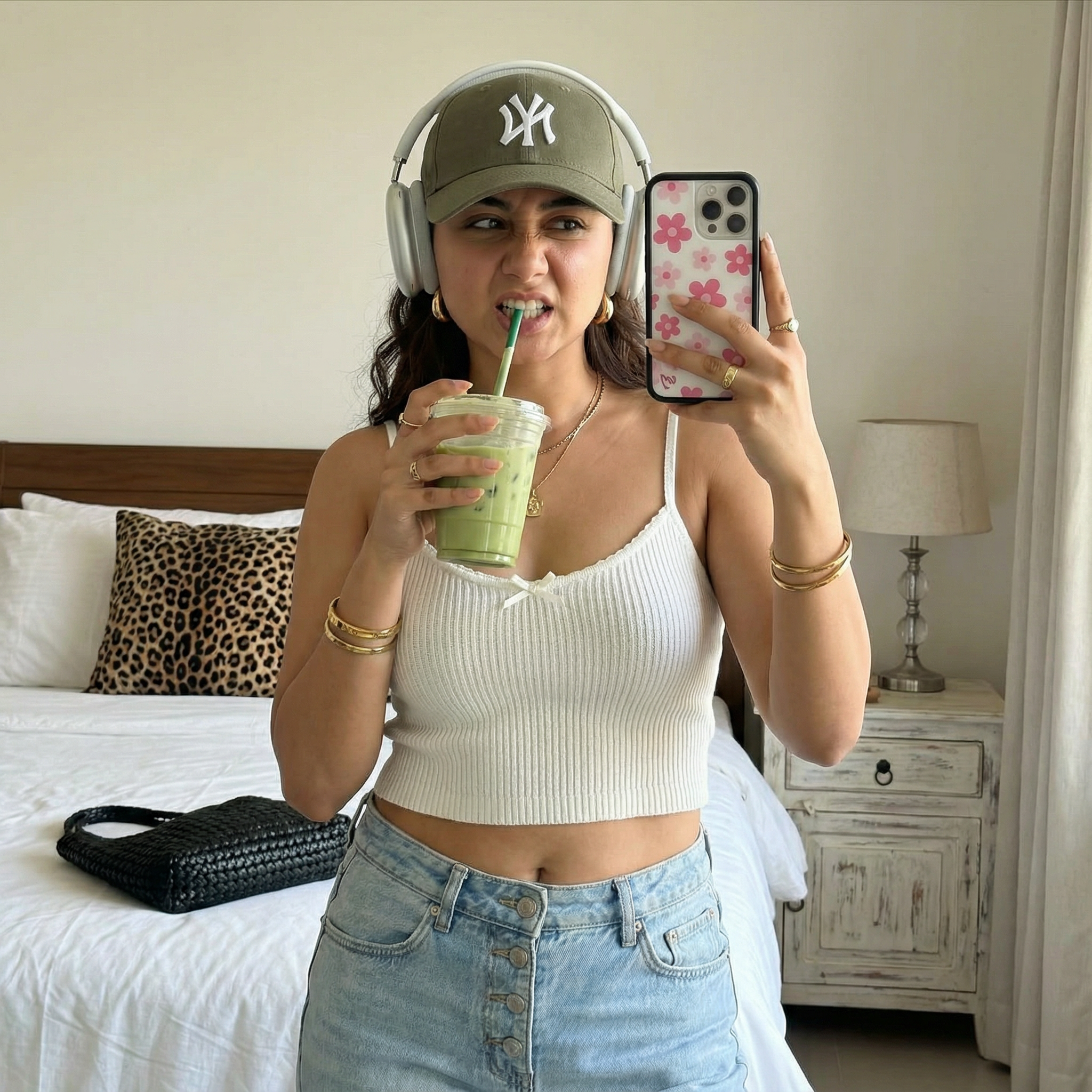 {
  "subject": {
    "description": "The person from the input image taking a mirror selfie, playfully interacting with the straw of an iced drink",
    "mirror_rules": "ignore mirror physics for text on clothing, display text forward and legible to viewer, no extra characters",
    "age": "match the person's real age",
    "expression": "playful, nose scrunched, gently biting or holding the straw",
    "hair": {
      "color": "match the person's real hair color",
      "style": "long straight hair or adapt to the person's natural hairstyle while keeping a similar overall vibe"
    },
    "clothing": {
      "top": {
        "type": "ribbed knit cami-style top (adapt fit appropriately to the person)",
        "color": "white",
        "details": "cropped fit, thin straps, small dainty bow at neckline"
      },
      "bottom": {
        "type": "denim jeans",
        "color": "light wash blue",
        "details": "relaxed fit, visible button fly"
      }
    },
    "face": {
      "preserve_original": true,
      "makeup": "natural sunkissed glow; if the person typically wears no makeup, apply only soft enhancement"
    }
  },
  "accessories": {
    "headwear": {
      "type": "olive green baseball cap",
      "details": "white NY logo embroidery, silver over-ear headphones worn over the cap"
    },
    "jewelry": {
      "earrings": "large gold hoops or adapt to the person's ear type",
      "necklace": "thin gold chain with small pendant",
      "wrist": "gold bangles and bracelets",
      "rings": "multiple gold rings"
    },
    "device": {
      "type": "smartphone",
      "details": "white case with pink floral pattern"
    },
    "prop": {
      "type": "iced beverage",
      "details": "plastic cup with iced matcha latte and green straw"
    }
  },
  "photography": {
    "camera_style": "smartphone mirror selfie aesthetic",
    "angle": "eye-level mirror reflection",
    "shot_type": "waist-up composition, subject positioned on the right side of the frame",
    "aspect_ratio": "9:16 vertical",
    "texture": "sharp focus, natural indoor lighting, social media realism, clean details"
  },
  "background": {
    "setting": "bright casual bedroom",
    "wall_color": "plain white",
    "elements": [
      "bed with white textured duvet",
      "black woven shoulder bag lying on bed",
      "leopard print throw pillow",
      "distressed white vintage nightstand",
      "modern bedside lamp with white shade"
    ],
    "atmosphere": "casual lifestyle, cozy, spontaneous",
    "lighting": "soft natural daylight"
  }
}