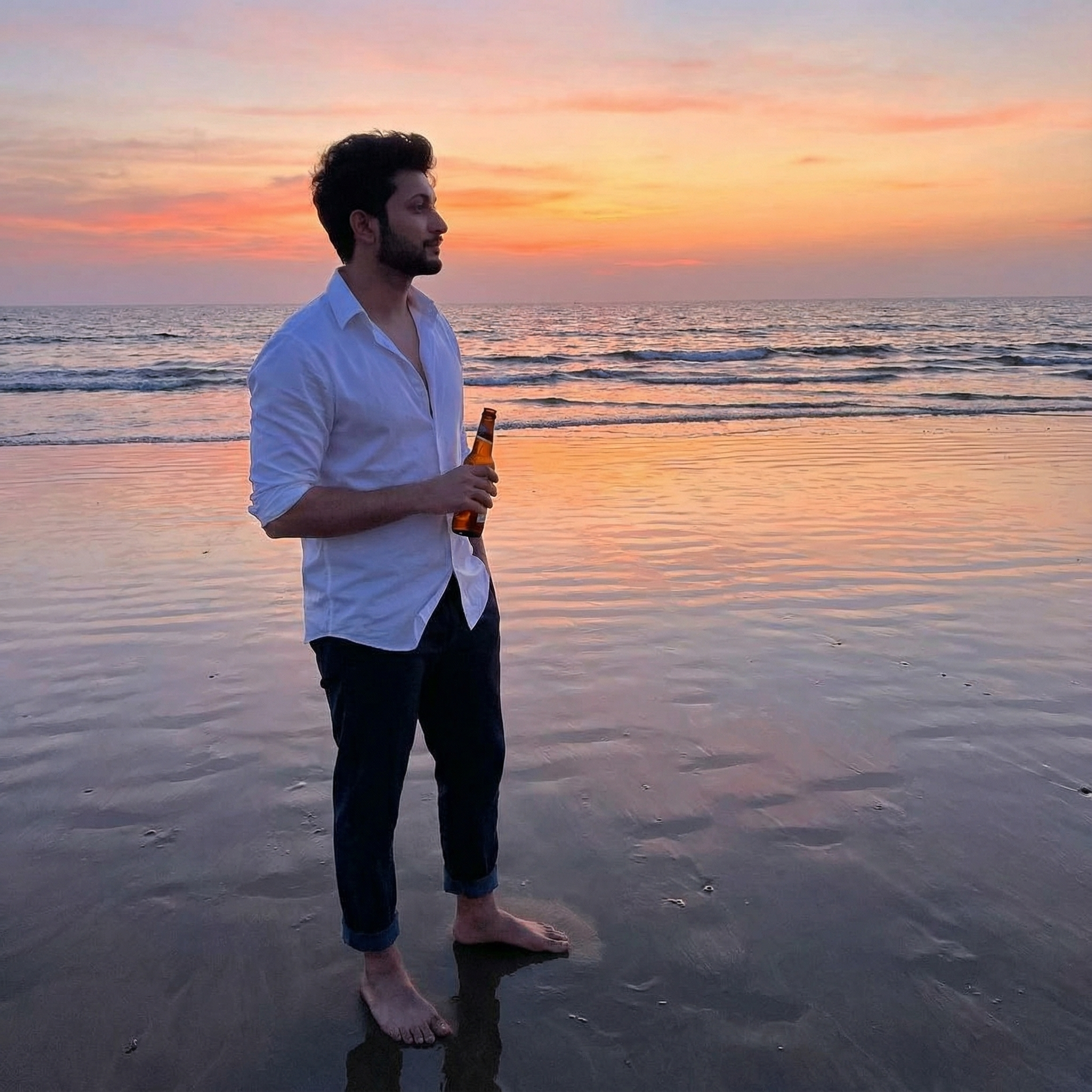 A candid shot standing on a beach at dusk, holding a beer bottle, watching the sun dip below the horizon. His trousers are rolled up, feet in wet sand. Lighting: Dramatic sunset colors reflecting on the wet sand.