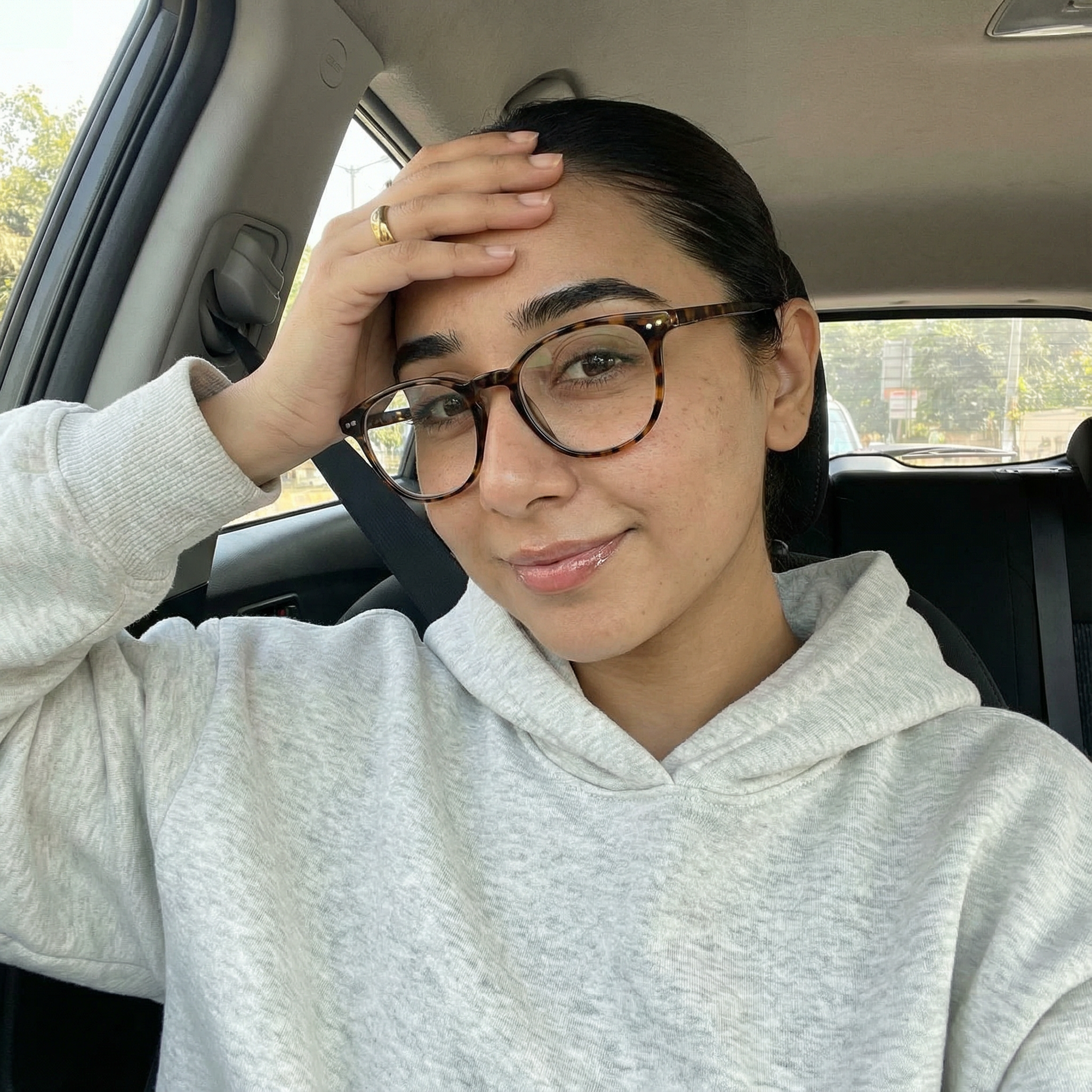 {
  "subject": {
    "description": "The person from the input image taking a car selfie with their hand resting on their forehead, smiling gently",
    "age": "match the person's real age",
    "expression": "relaxed, candid, slight smile, hand casually touching or resting on the forehead",
    "hair": {
      "color": "match the person's natural hair color",
      "style": "slicked-back tight low bun with a precise middle part, or adapt naturally to the person's real hairstyle"
    },
    "clothing": {
      "top": {
        "type": "oversized hoodie",
        "color": "light heather grey",
        "details": "soft fleece fabric, relaxed fit, hood resting back"
      }
    },
    "face": {
      "preserve_original": true,
      "makeup": "fresh natural 'clean aesthetic'; if the person does not typically wear makeup, apply only soft enhancements like sun-kissed skin, subtle freckles, light blush, natural glossy lips, and groomed brows"
    }
  },
  "accessories": {
    "eyewear": {
      "type": "tortoise-shell glasses",
      "details": "oval or round acetate frames"
    },
    "earrings": {
      "type": "gold ear stack",
      "details": "multiple small gold huggie hoops and studs on lobe and helix"
    },
    "jewelry": {
      "necklace": "dainty gold chain with a tiny pendant",
      "ring": "thin gold band on the ring finger"
    }
  },
  "photography": {
    "camera_style": "modern smartphone selfie",
    "angle": "eye-level or slightly low angle",
    "shot_type": "close-up portrait",
    "aspect_ratio": "9:16 vertical",
    "texture": "natural daylight, sharp focus on the face, soft realistic skin texture, bright window lighting, no grain"
  },
  "background": {
    "setting": "interior of a car",
    "elements": [
      "dark car ceiling or panoramic sunroof",
      "car seat headrest",
      "seatbelt",
      "car window showing bright daylight",
      "blurred trees and buildings outside"
    ],
    "atmosphere": "casual daily life, relaxed, bright daytime mood",
    "lighting": "soft natural window light illuminating the face"
  }
}