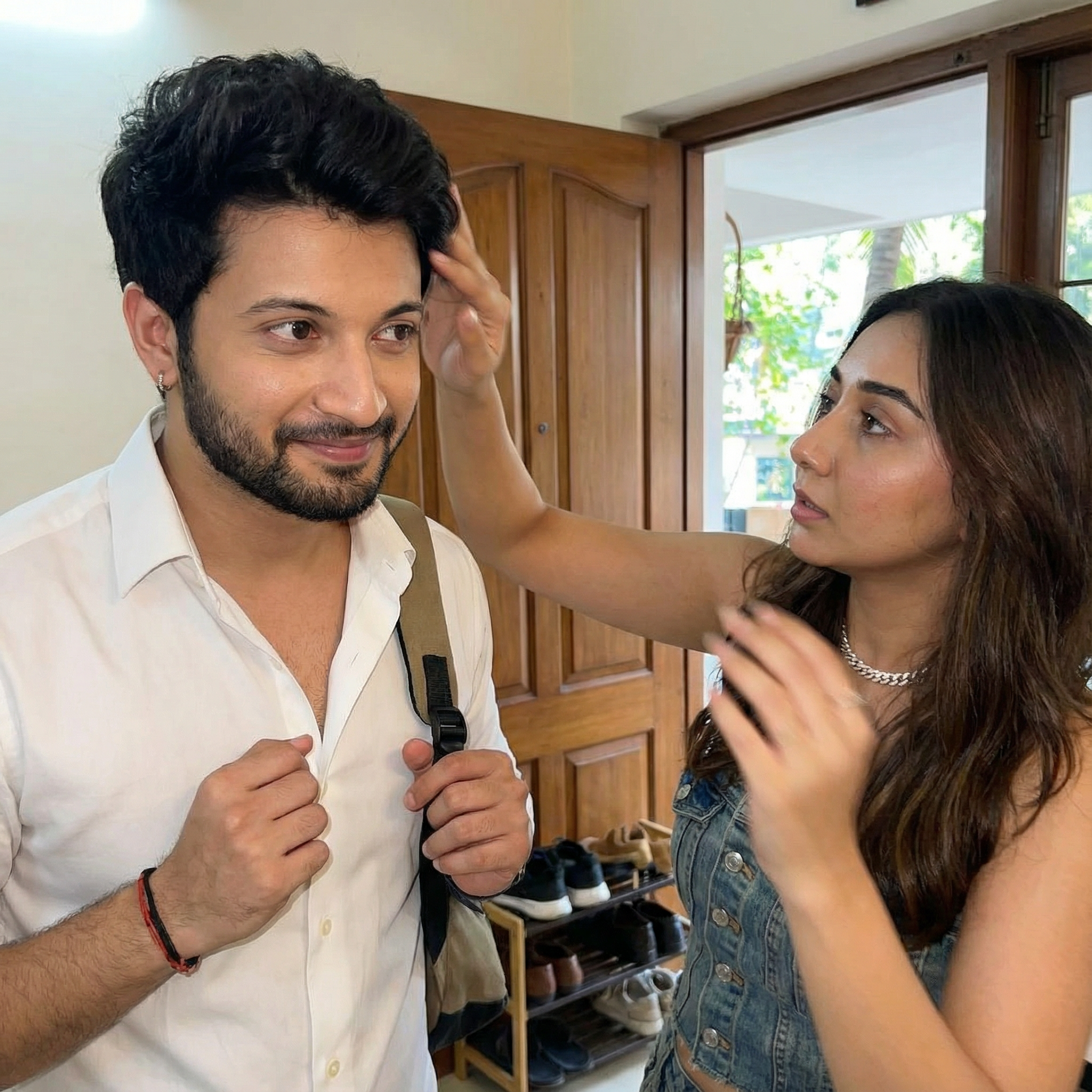 A close-up, fast-paced shot of an Indian couple at their front door. The man is looking slightly impatient to leave, while the girl is quickly adjusting his shirt collar or smoothing down his hair. Her expression is focused and caring, and his is appreciative, capturing the quick, close intimacy of commute prep.
