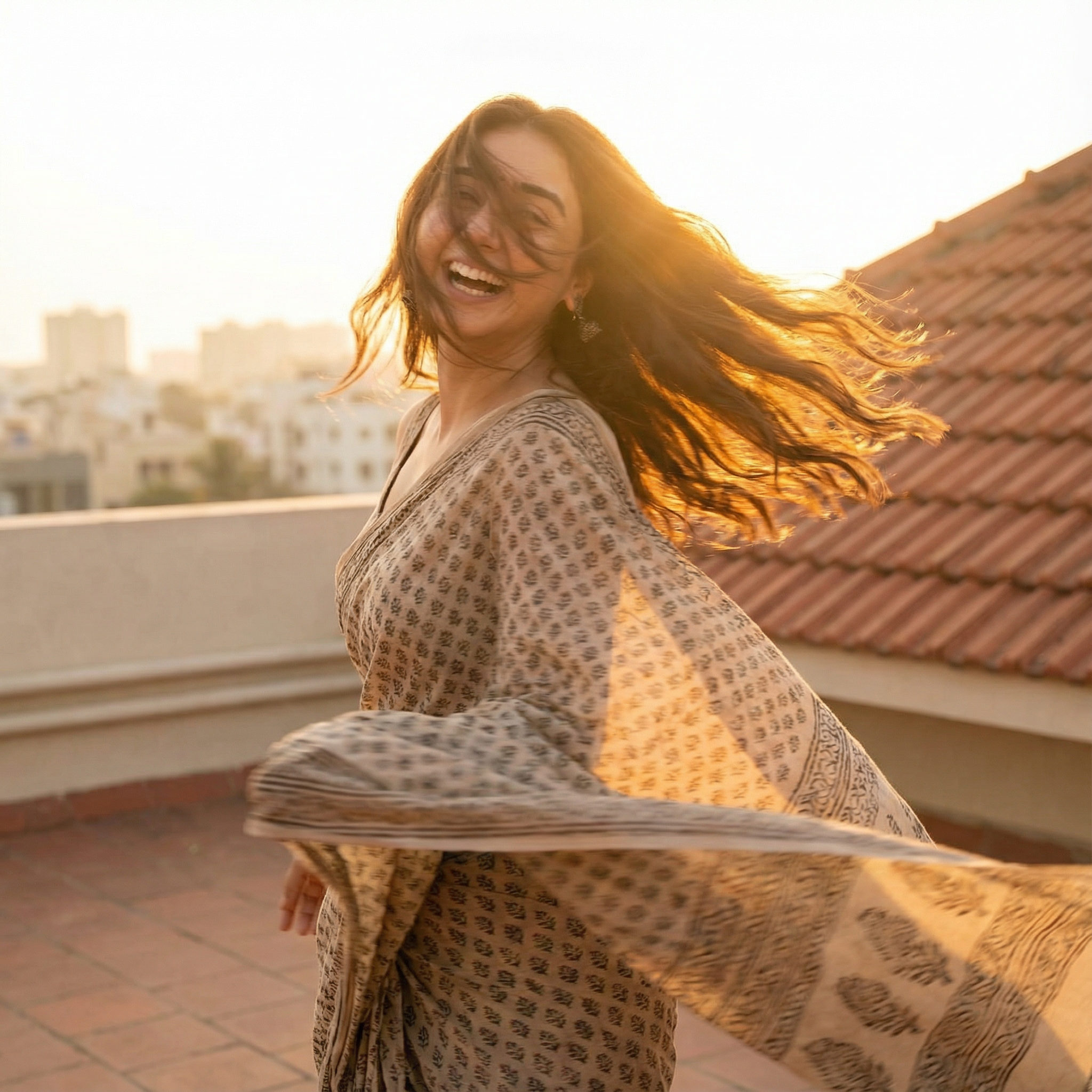 A candid, motion-blurred shot of a girl spinning around in a simple cotton saree on a rooftop terrace at sunset. She is looking back over her shoulder. Expression Cue: Her hair is flying across her face. She is laughing uncontrollably, looking breathless and free. Not a posed model smile. Lighting: Golden hour "halo" backlight.