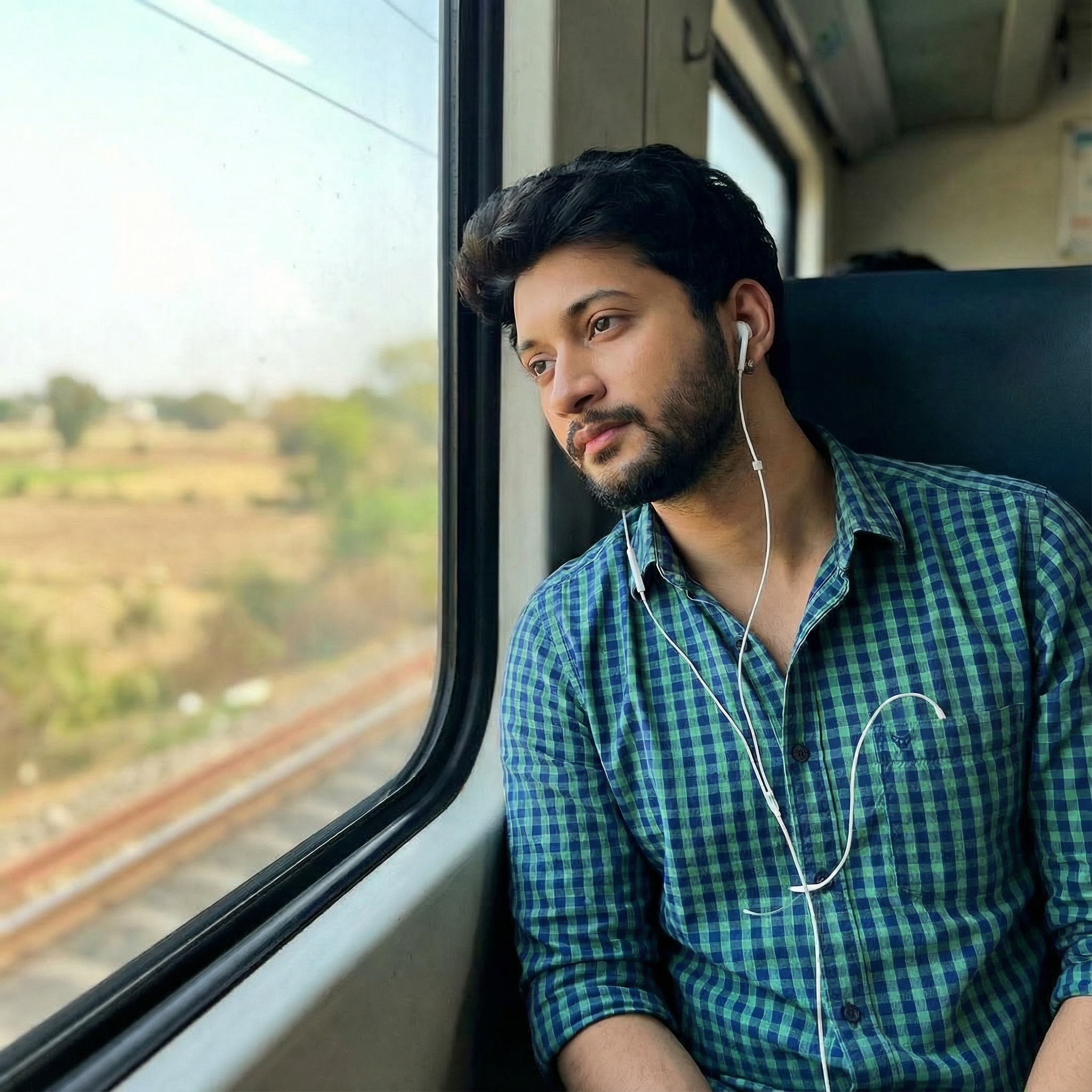 Sitting by the window seat of an Indian Railways train, resting his head against the glass,wearing white wired earphones, watching the landscape blur past. He looks thoughtful, wearing a simple checked shirt. Lighting: Natural daylight from the window.