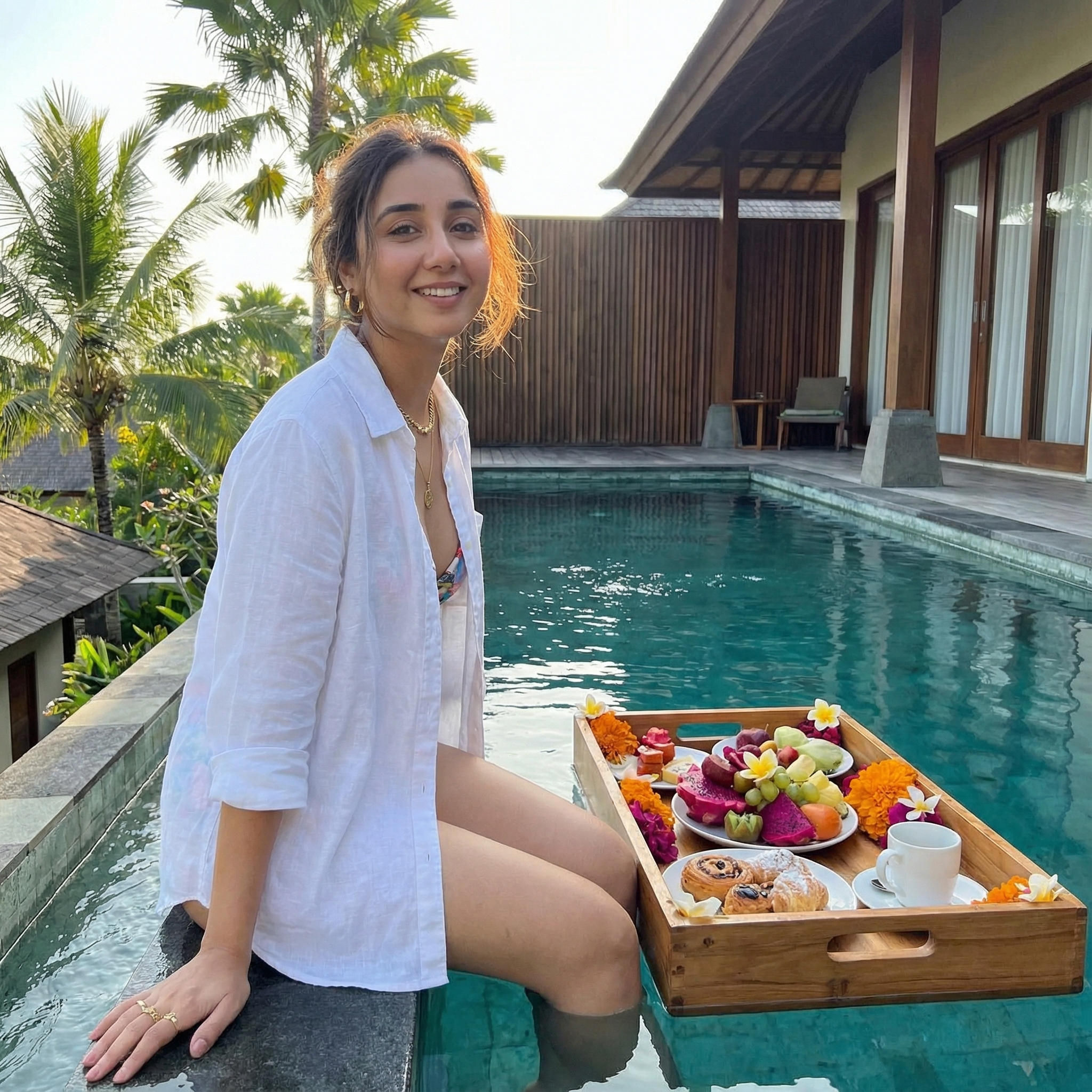 {
  "subject": {
    "description": "The person from the input image sitting beside a crystal-blue pool with a floating breakfast tray",
    "expression": "relaxed, warm, peaceful luxury vacation mood",
    "hair": {
      "style": "natural tropical styling, soft texture"
    },
    "clothing": {
      "outfit": "white linen resort wear or luxury swimwear",
      "details": "light airy fabric movement, sunlit highlights"
    }
  },
  "accessories": {
    "prop": "floating tray filled with tropical fruits, pastries, flowers",
    "jewelry": "minimal gold pieces"
  },
  "photography": {
    "camera_style": "tropical luxury influencer aesthetic",
    "angle": "slightly elevated poolside",
    "shot_type": "mid-shot",
    "texture": "vibrant colors, soft sunlight, lush clarity"
  },
  "background": {
    "setting": "Bali private villa",
    "elements": [
      "infinity pool",
      "palm trees",
      "wooden villa architecture",
      "tropical flowers and shadows"
    ],
    "atmosphere": "serene tropical wealth",
    "lighting": "bright soft morning sunlight"
  }
}