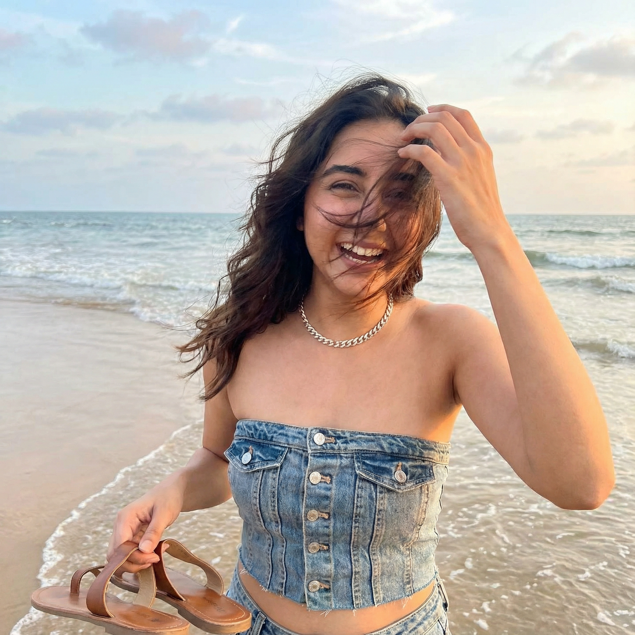 A girl standing on a beach with her feet in the water, holding her sandals in one hand. The wind is blowing her hair completely over her face. Expression Cue: She is brushing the hair away with a hand, laughing at the wind. Not a perfect hair flip—a messy, real, happy moment.